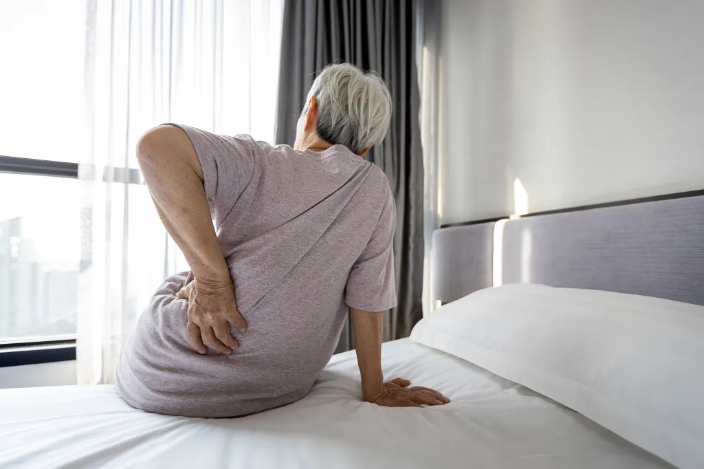 An older Asian woman sits on a bed, massaging her lower back due to pain.