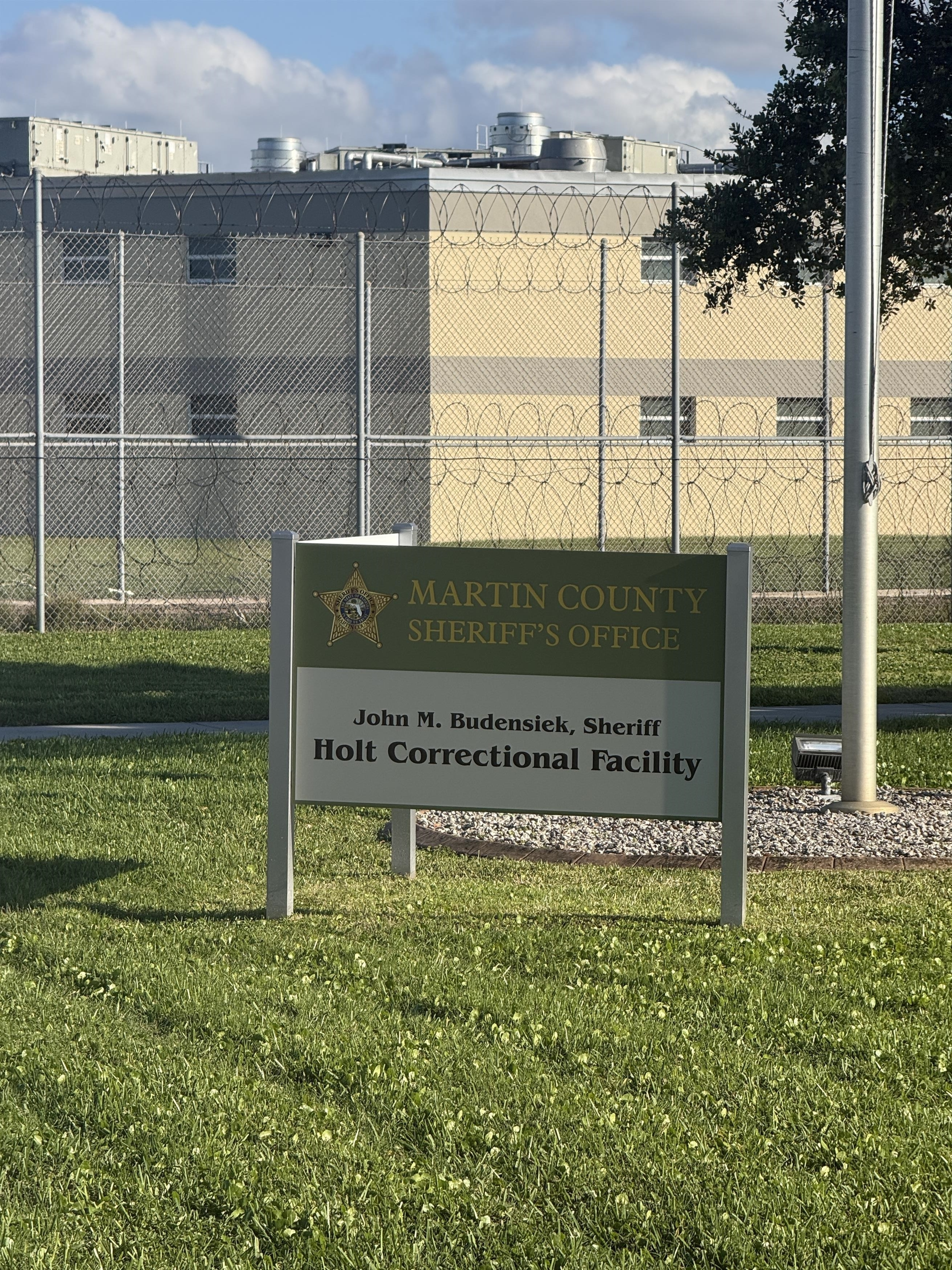 Sign for the Martin County Sheriff's Office Holt Correctional Facility, with the facility behind a fence topped with barbed wire.