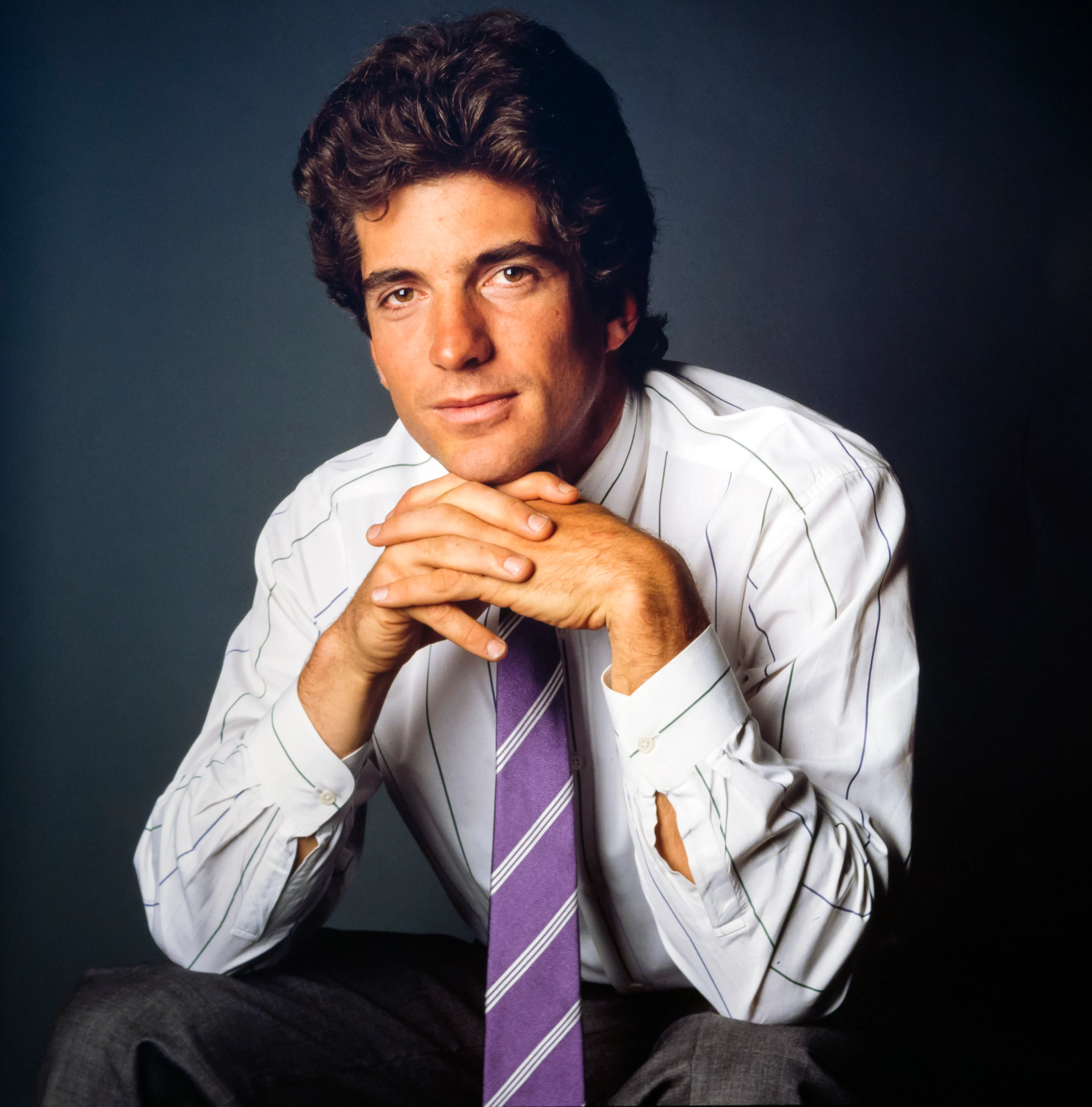 Studio portrait of American lawyer and magazine publisher John F. Kennedy Jr., wearing a white shirt and purple striped tie, with his hands clasped under his chin.