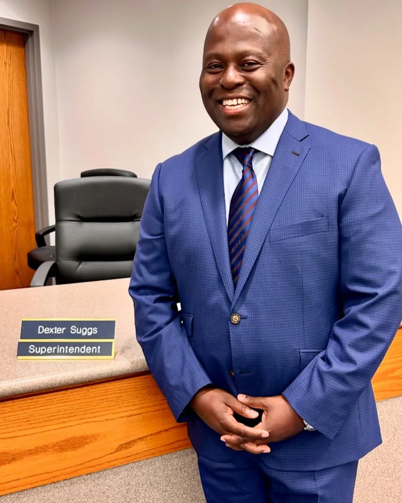 Superintendent Dexter Suggs smiling while standing next to a desk with a nametag that reads 