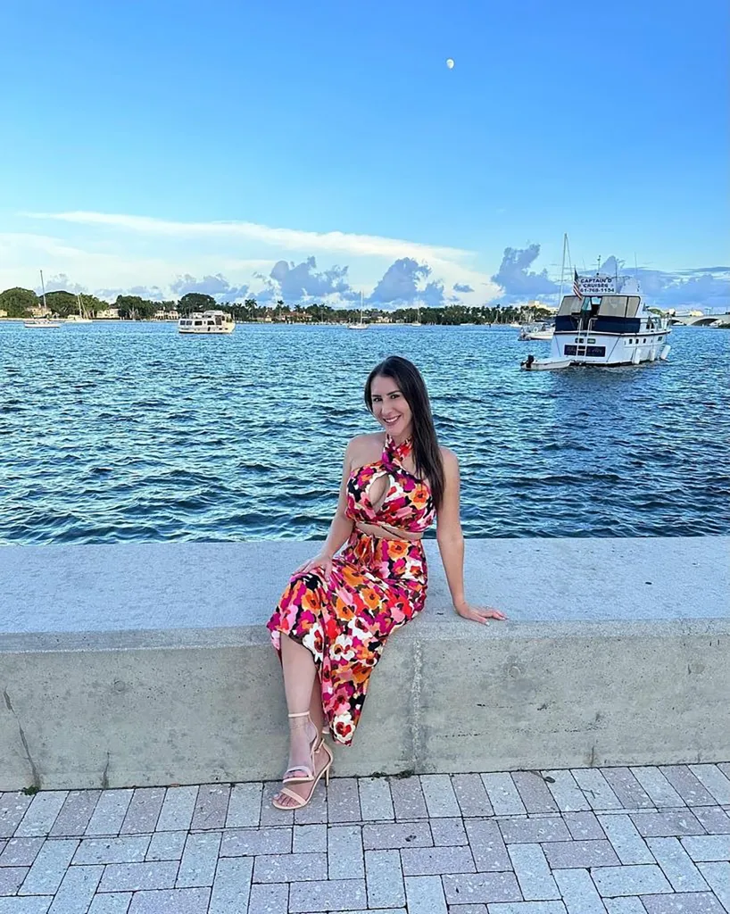 A woman in a floral dress and heels sitting by the water with boats in the background.
