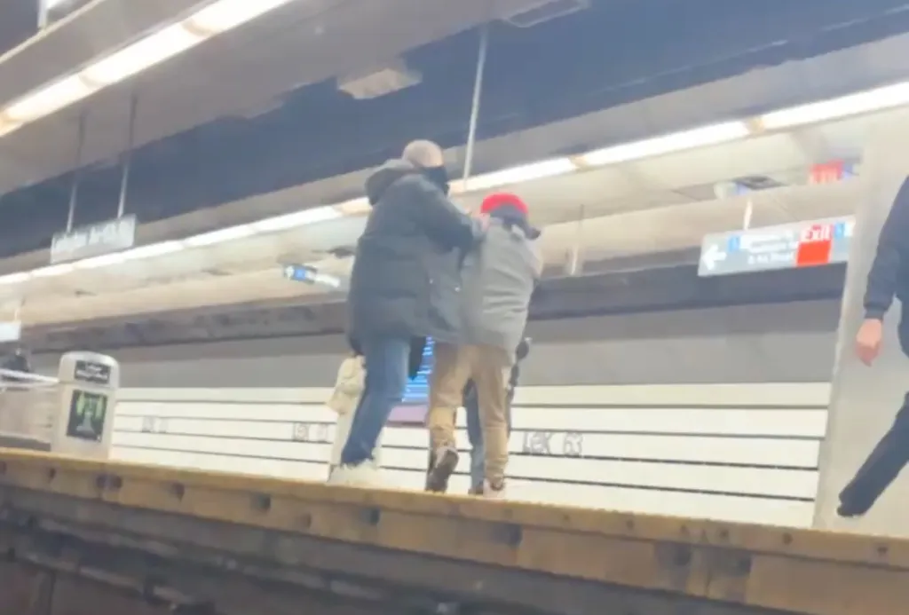 Two men on a subway platform, one in a black coat and the other in a red hoodie and beige pants, with the man in the black coat seemingly holding the man in the red hoodie from behind.