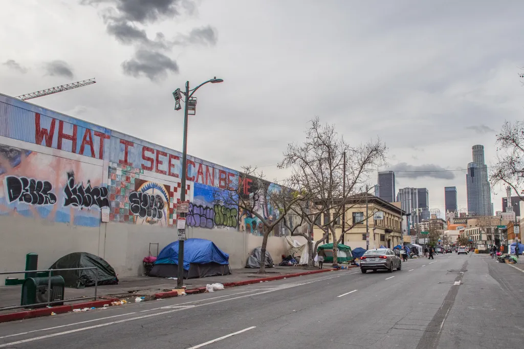 Tents line the street in Skid Row, Los Angeles, with graffiti on a wall and skyscrapers in the background.