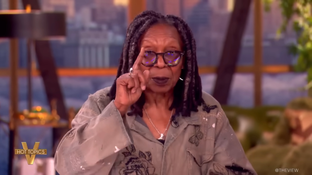Whoopi Goldberg points her finger, wearing an olive green jacket, glasses, and a necklace.