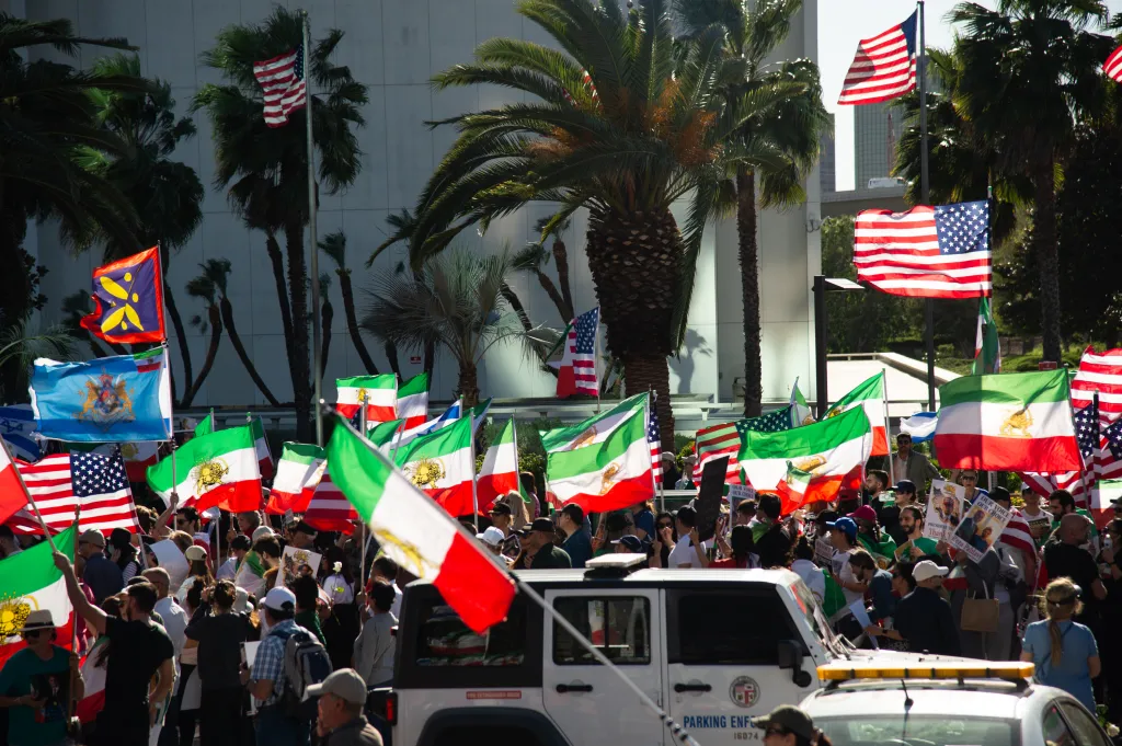 Thousands of people gathered in Westwood, CA, holding up Iranian and American flags, with palm trees and a building in the background.