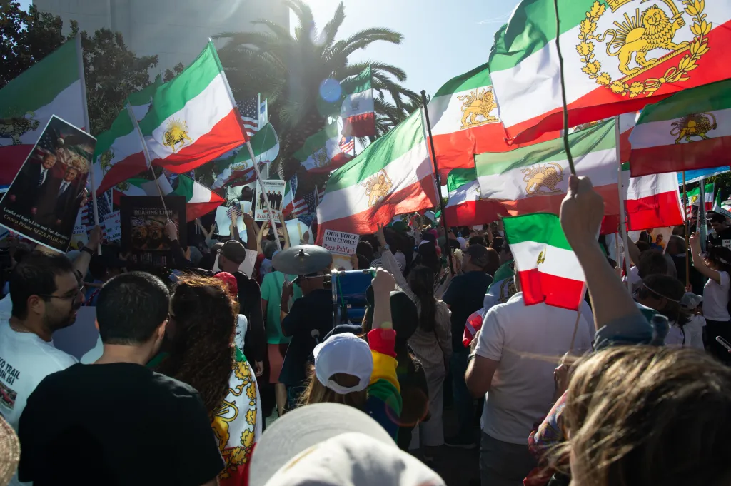 Crowd of people at a rally holding Iranian flags with the lion and sun emblem, and signs supporting Reza Pahlavi.