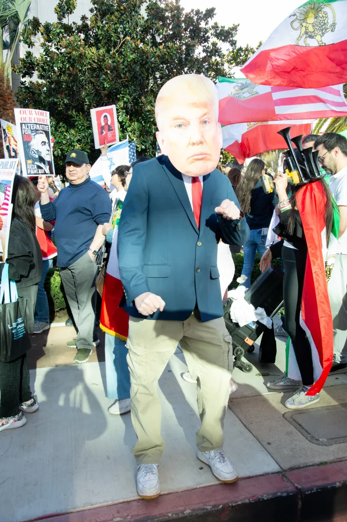 A man wearing a Donald Trump mask and suit jacket stands among a crowd carrying Iranian flags and protest signs.