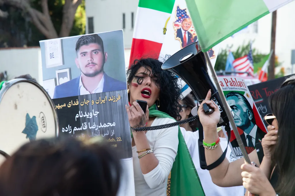 A woman speaking into a megaphone at a protest, surrounded by signs including a portrait of a young man, a flag of Iran with the sun and lion emblem, a banner of former President Trump, and a poster of the former Shah of Iran, Reza Pahlavi.