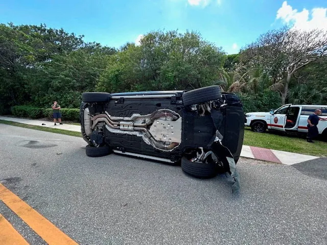 An overturned black Land Rover lies on its side in the middle of a two-lane road.