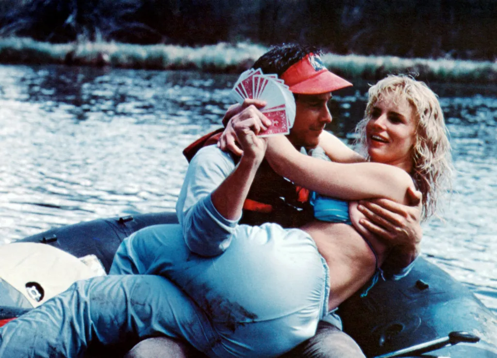 Tim Matheson holding a fan of playing cards and Jennifer Runyon in a blue bikini embrace in an inflatable raft.