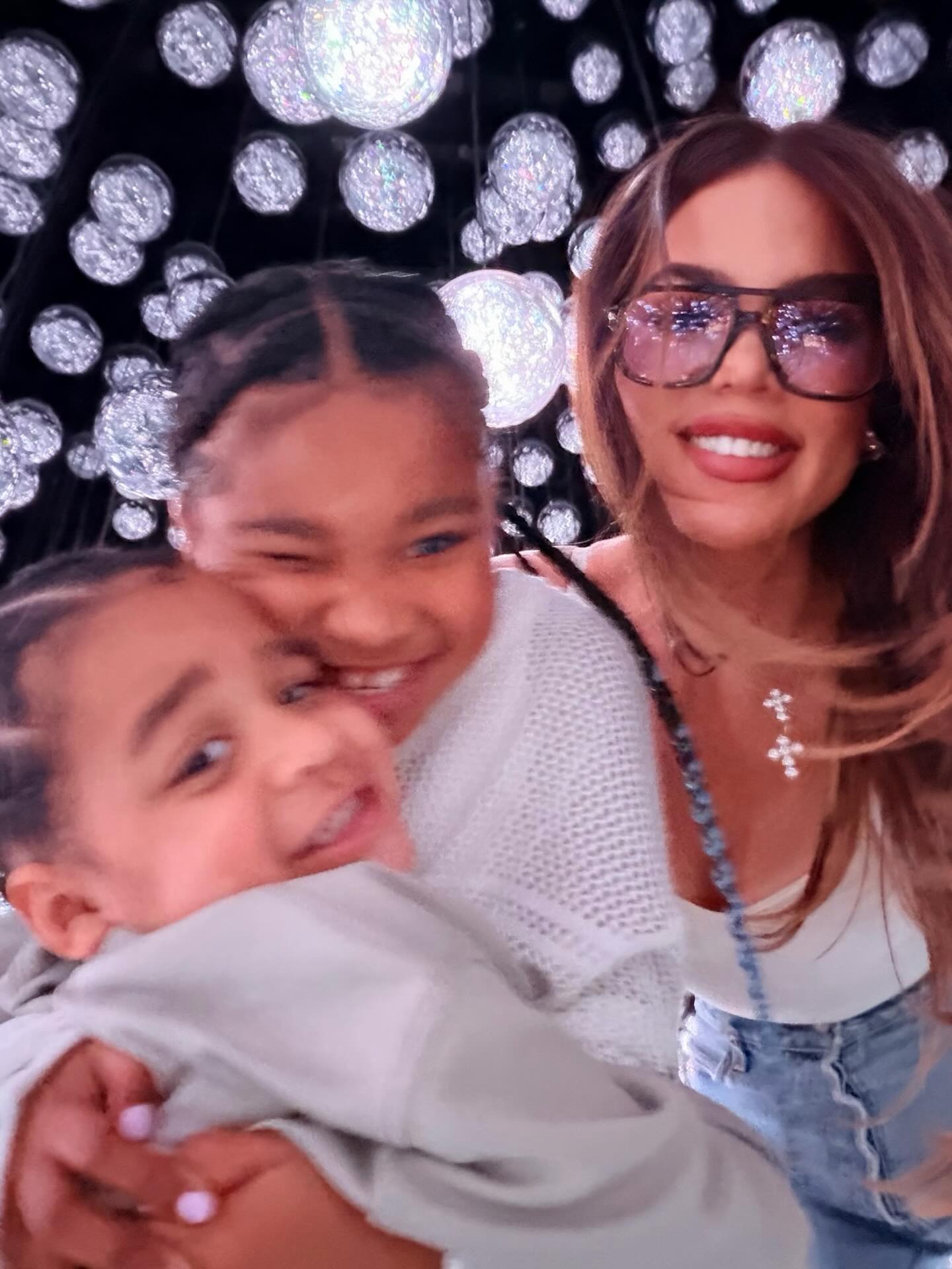 Khloé Kardashian with her two children, True and Tatum, posing in front of a sparkling light display.