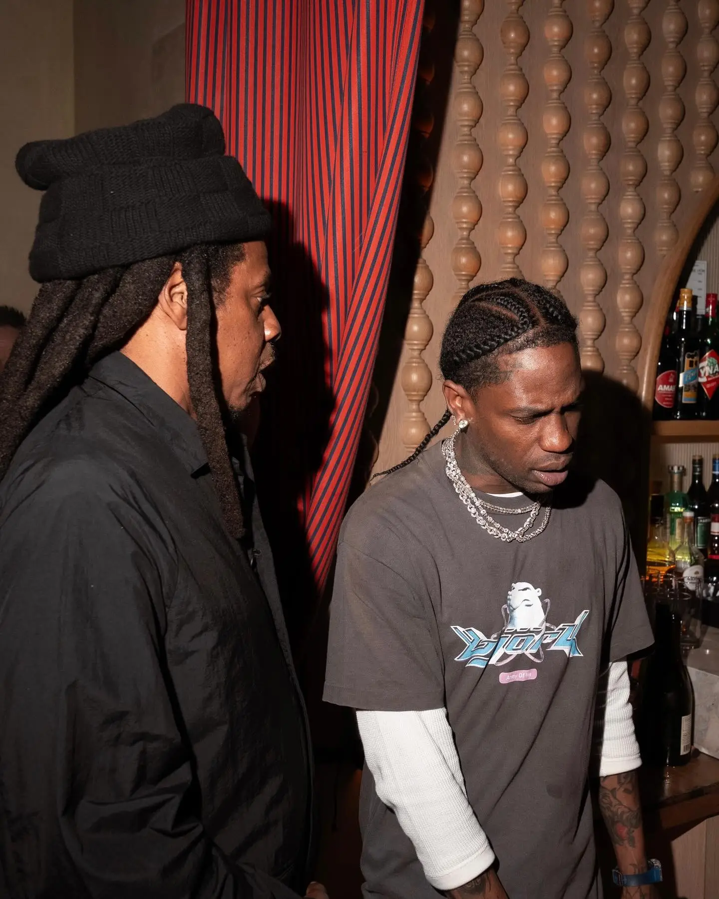 Jay-Z (left) in a black jacket and beanie, and Travis Scott (right) in a gray t-shirt and white undershirt, talking at a Fanatics Studios bash.