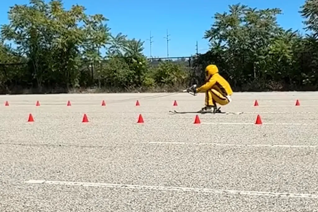 Man in a yellow suit on skis on an asphalt parking lot between orange cones.