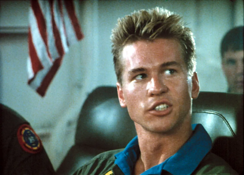 Val Kilmer as Iceman in a military classroom, wearing a flight suit, with an American flag in the background.