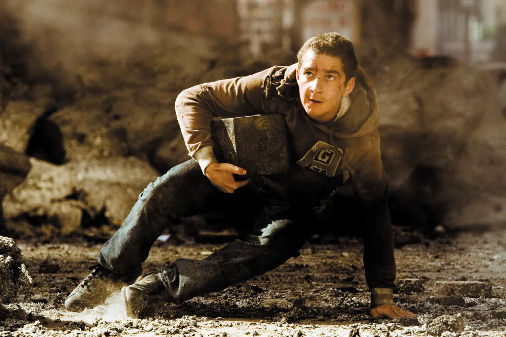 Shia LaBeouf, in a dark hoodie and black pants, as Sam Witwicky in Transformers, holding the AllSpark cube, looking up with a scared expression while on the ground.
