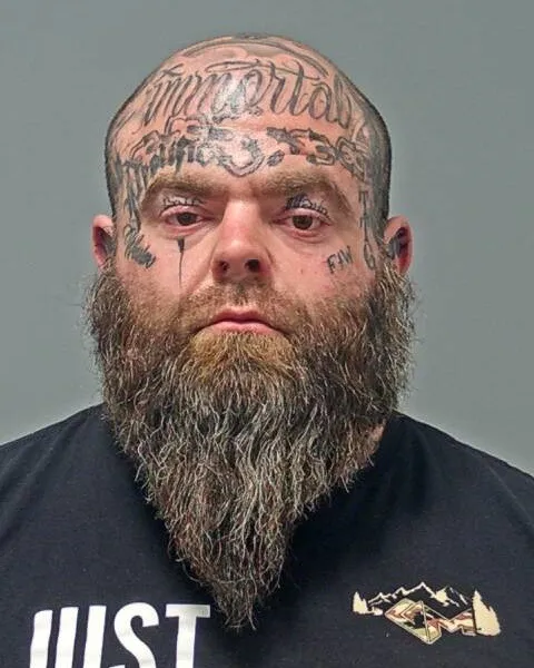 Mugshot of a man with face and head tattoos and a long beard.