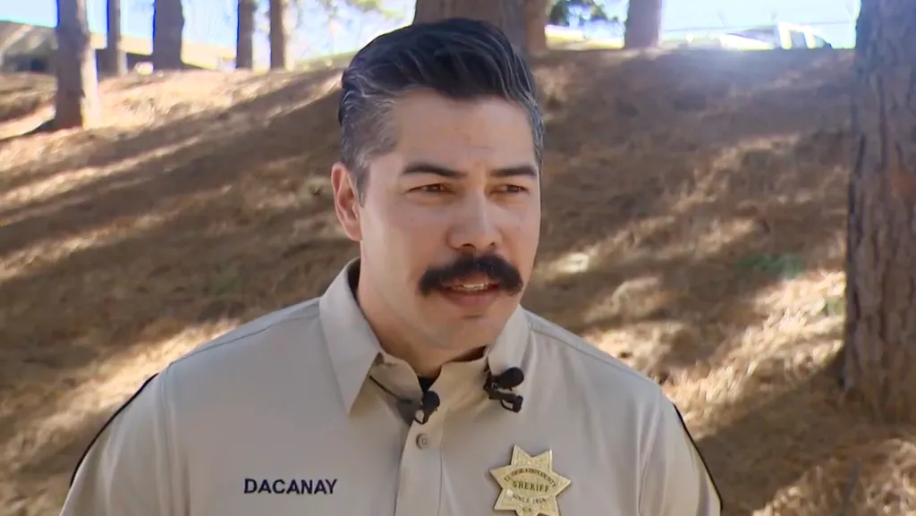 A sheriff deputy with a mustache and badge speaks to the camera.