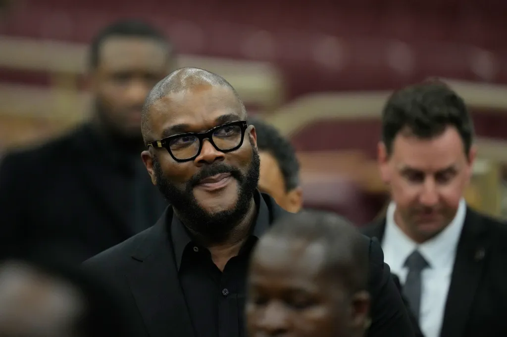 Tyler Perry wearing black-framed glasses, a black suit jacket, and a black shirt.