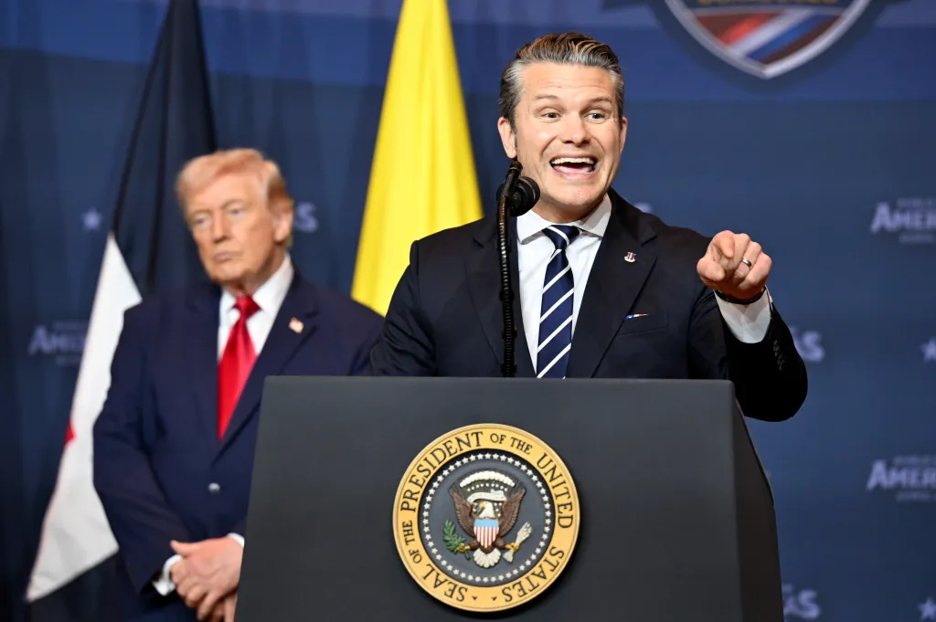 U.S. Defense Secretary Pete Hegseth speaks from a podium with the Seal of the President of the United States, while President Donald Trump stands behind him.
