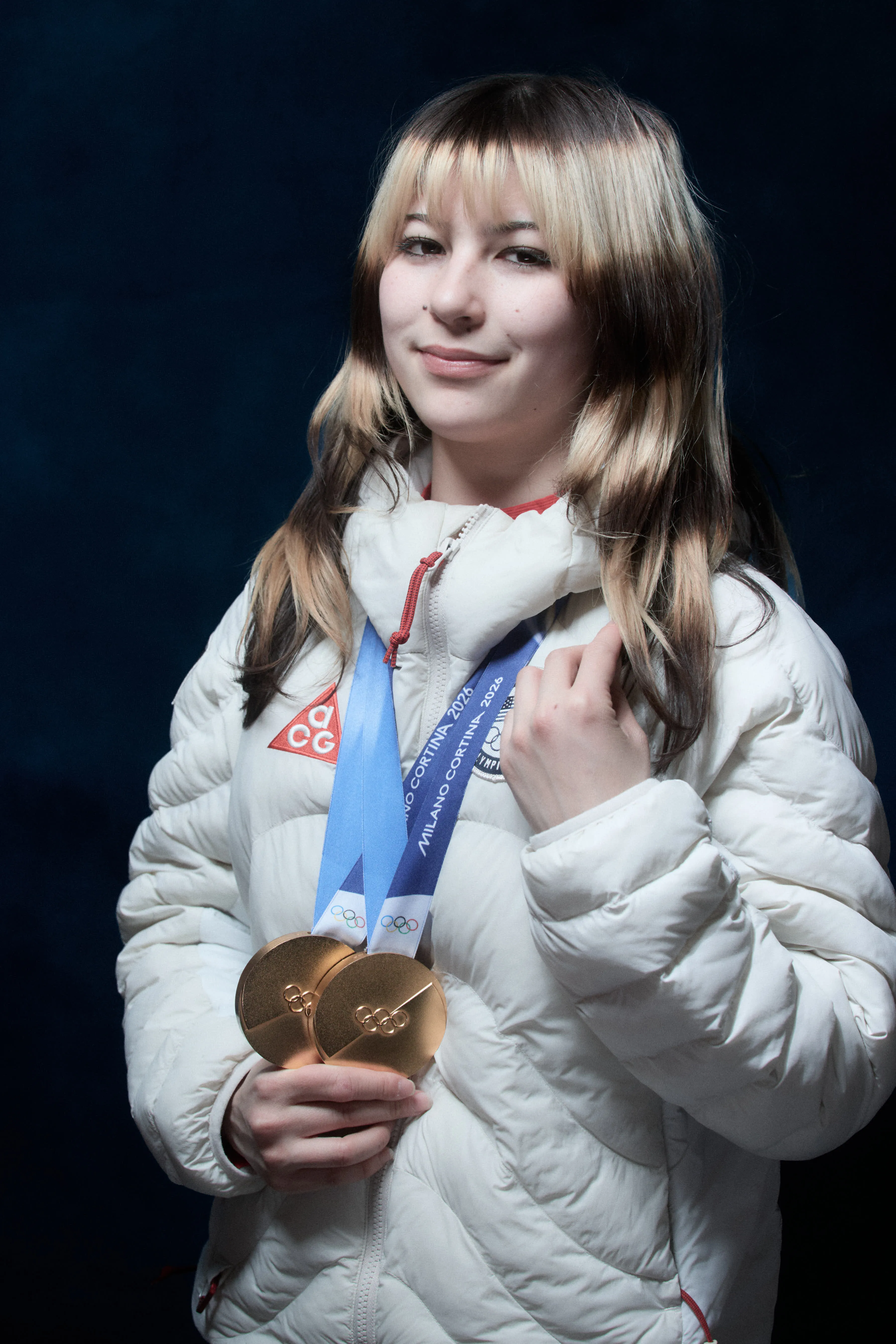Alysa Liu, a US Olympian, wearing a white puffy jacket and holding two gold medals around her neck.