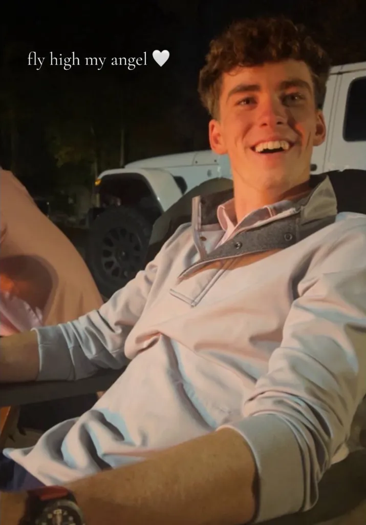 A young man with curly brown hair and a wide smile, wearing a gray collared shirt, sits in a chair with a white jeep visible in the background.