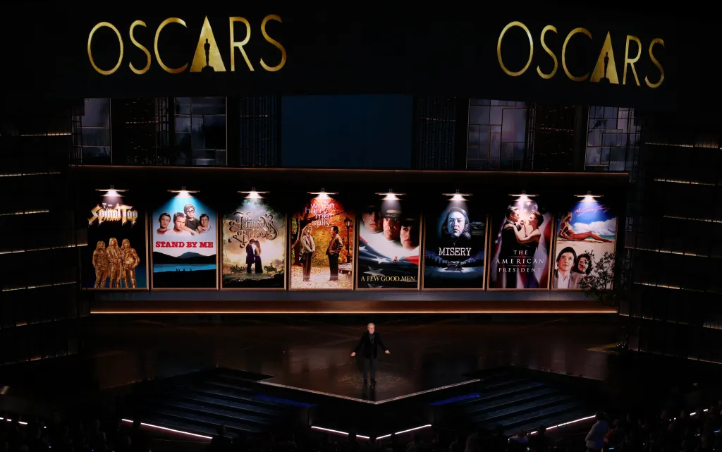 Billy Crystal onstage at the Oscars, standing before a screen displaying movie posters from director Rob Reiner.