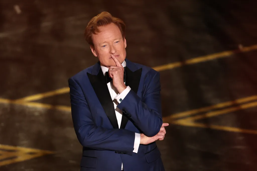 Conan O'Brien onstage at the Academy Awards, wearing a blue tuxedo, with his finger to his lips.