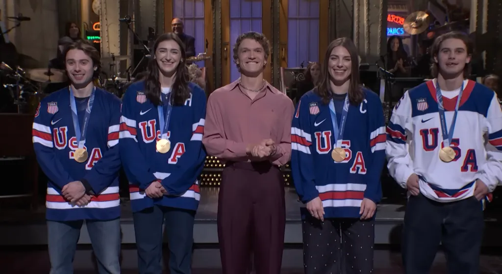 Connor Storrie on SNL with Quinn Hughes, Jack Hughes, Hilary Knight, and Megan Keller, all wearing USA hockey jerseys and medals.