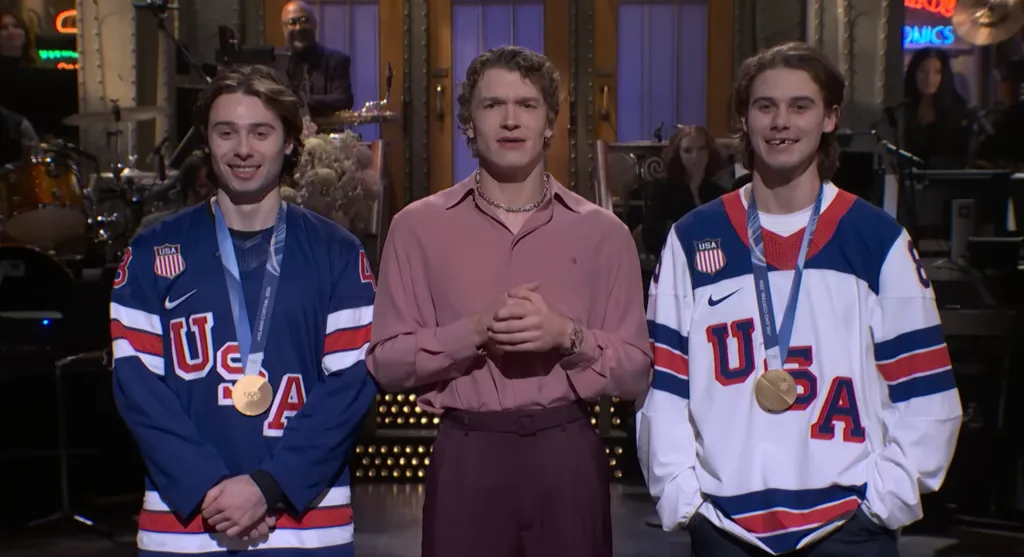 Connor Storrie stands between Quinn Hughes and Jack Hughes, who wear Team USA jerseys with gold medals and a missing front tooth on Jack.