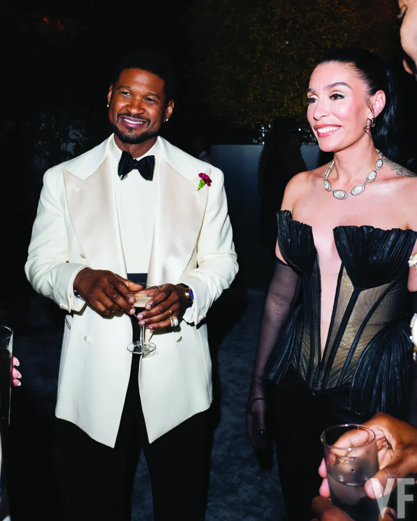 Usher, in a white tuxedo, smiling and holding a cocktail while standing next to a woman in a black dress at an after-party.