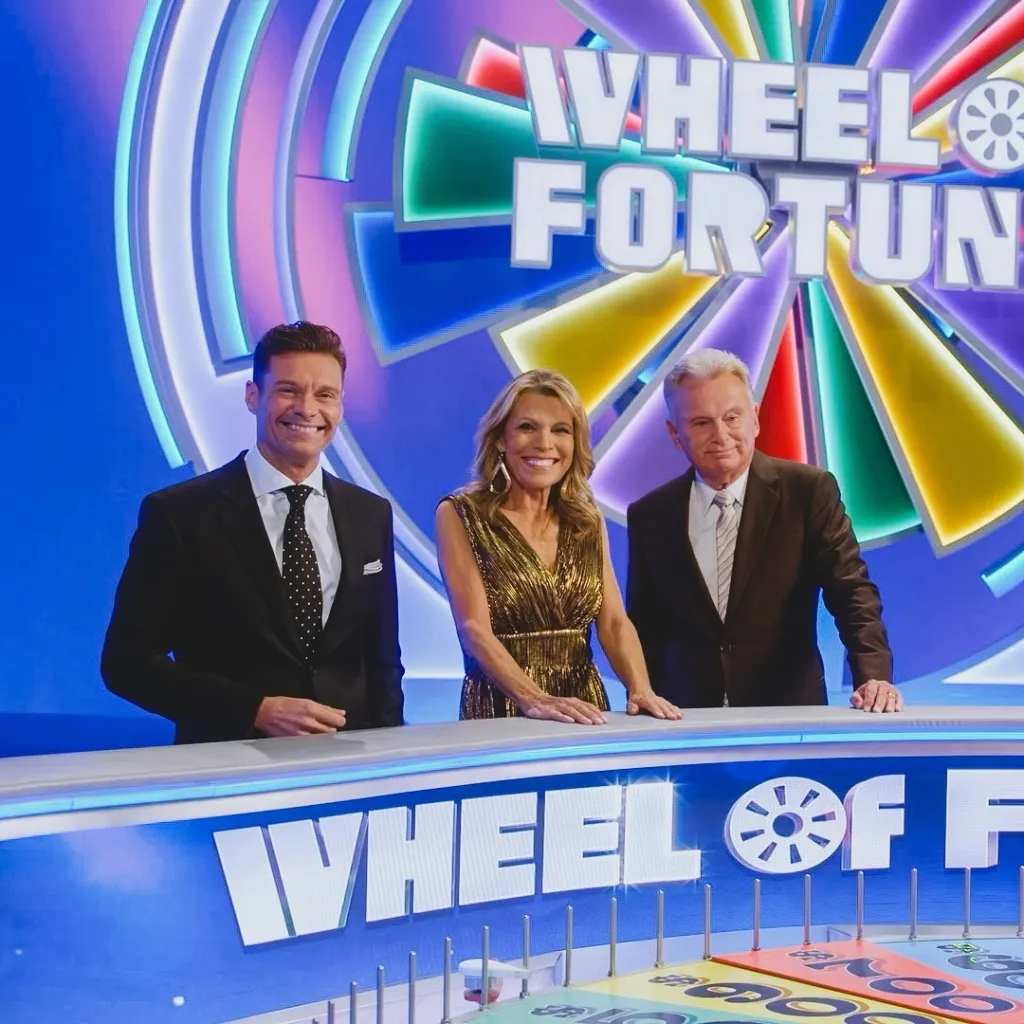 Ryan Seacrest, Vanna White, and Pat Sajak standing together on the Wheel of Fortune set.