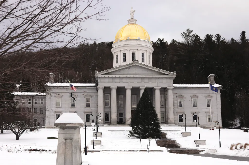 The Vermont State House, a neoclassical building with a golden dome and a statue on top, stands covered in snow with a forest behind it.