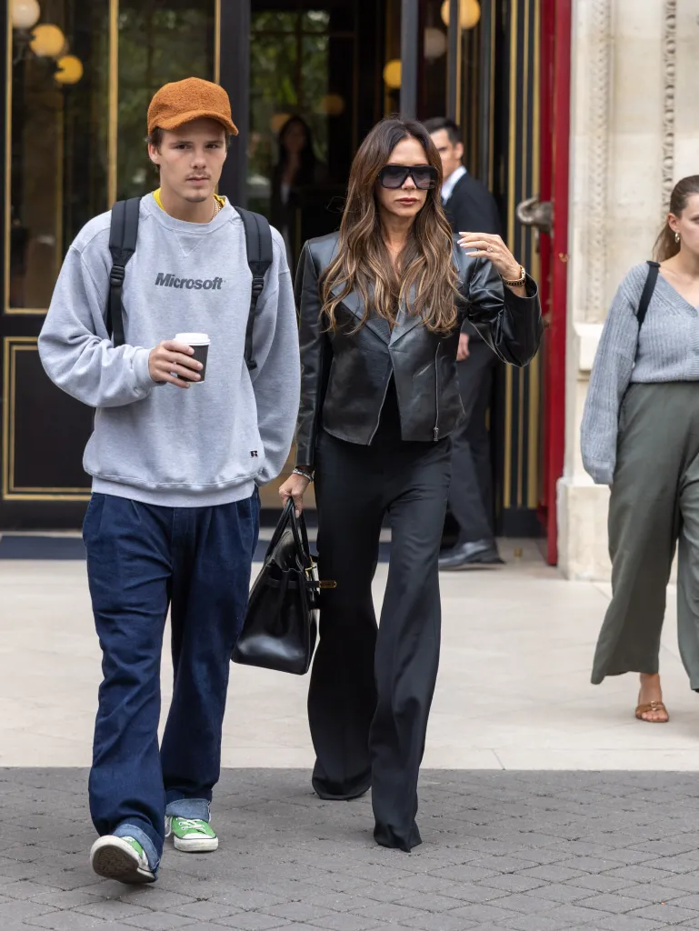 Victoria Beckham in a black leather jacket and sunglasses walks beside Cruz Beckham wearing a Microsoft sweatshirt and a brown cap.