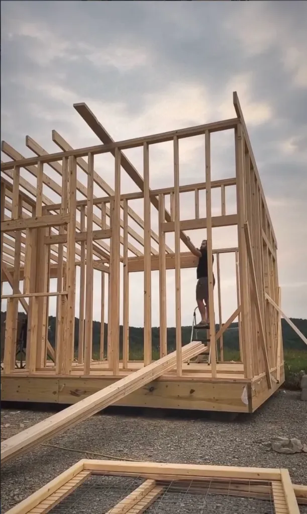 Tyler Neidhardt standing on a ladder, working on the wooden frame of a tiny house.