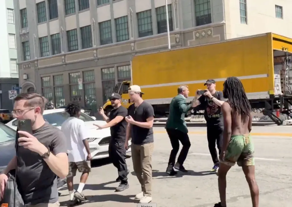 People filming a street confrontation in front of a yellow moving truck.