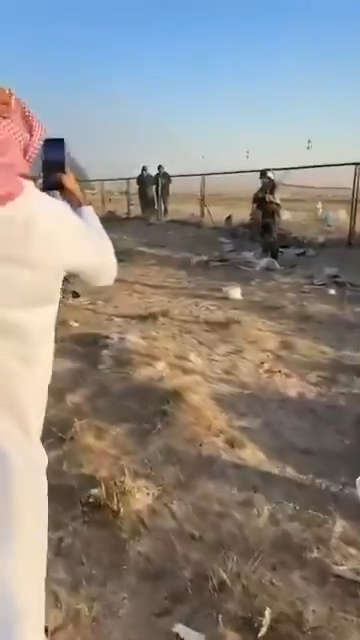 A man in a flight suit stands next to a fence in a desert landscape as bystanders look on.