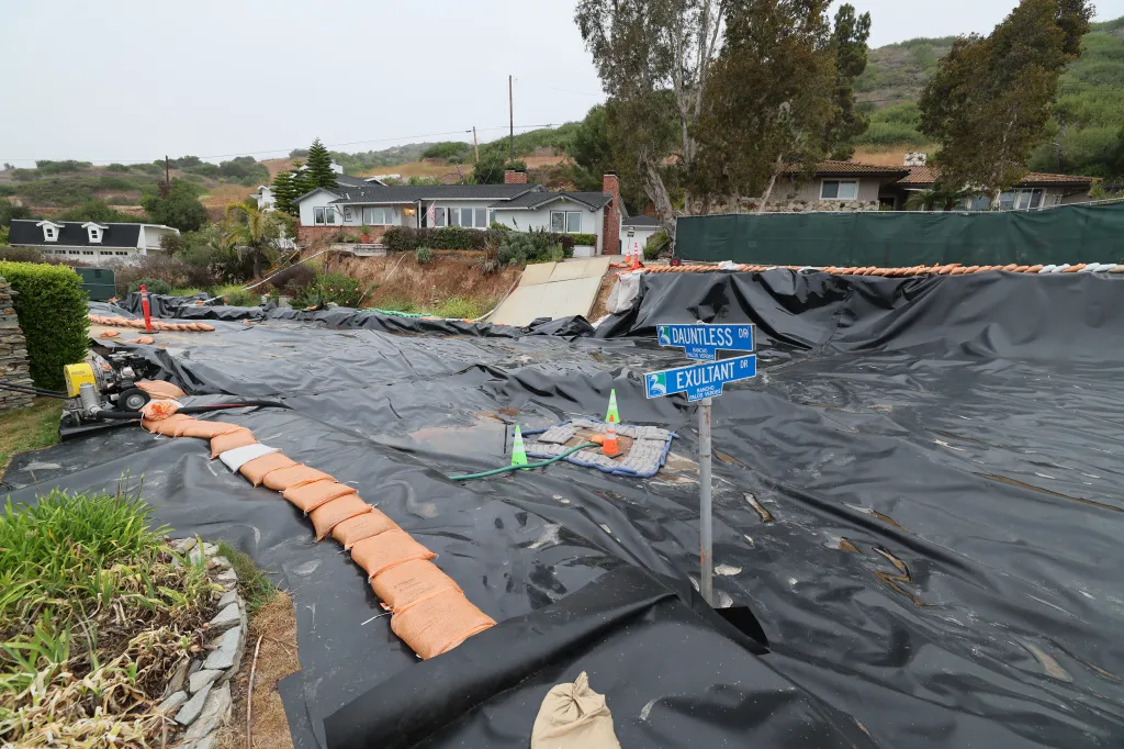 Landslide damage in Rancho Palos Verdes, California, showing houses on a damaged hillside, covered in black tarps and sandbags.