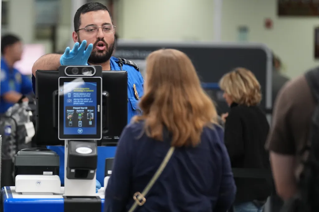 A TSA agent wearing gloves directs a traveler through a security checkpoint with a facial recognition system.