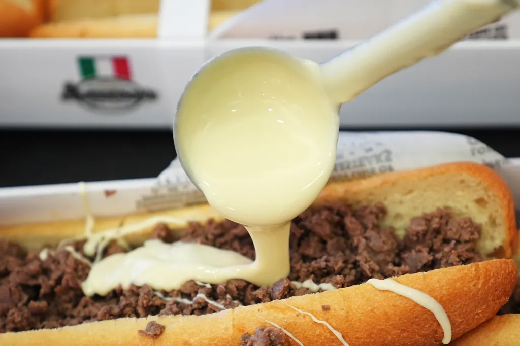 A ladle pours cheese sauce over a long cheesesteak in a white tray.