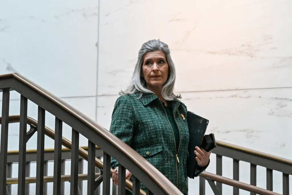 Senator Joni Ernst descends stairs, holding a folder and a device, likely on her way to a Senate Armed Services Committee briefing.