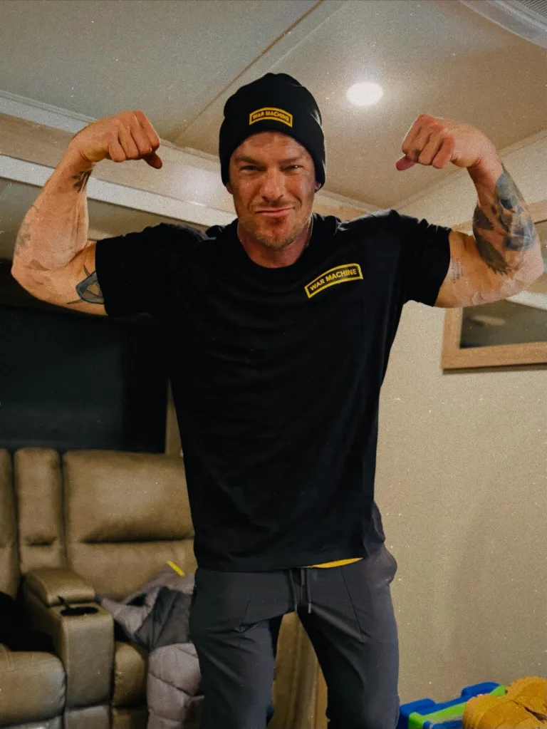 Alan Ritchson flexing his biceps while wearing a black T-shirt and beanie with 