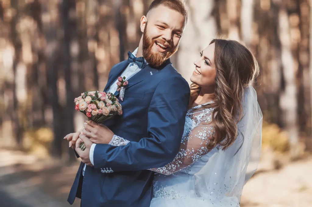 A smiling groom holds his bride close as she looks up at him, both happy in a park.