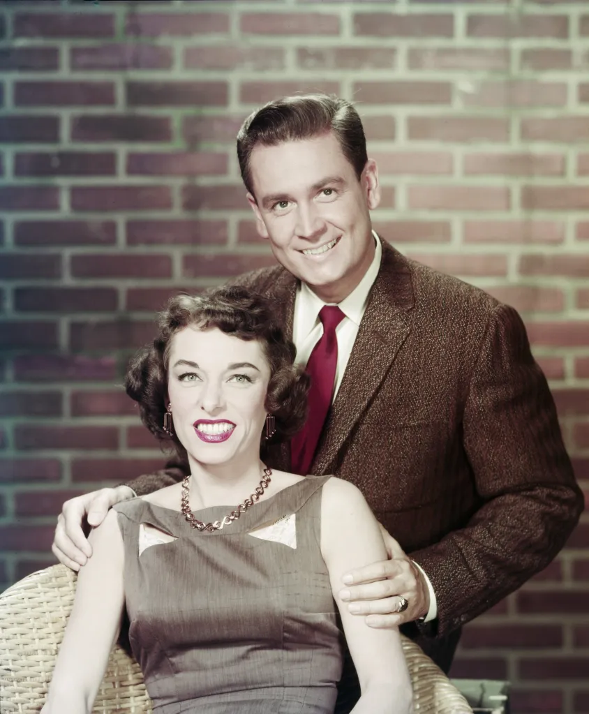 Bob Barker stands smiling behind his wife, Dorothy Jo Barker, who is seated in a wicker chair.