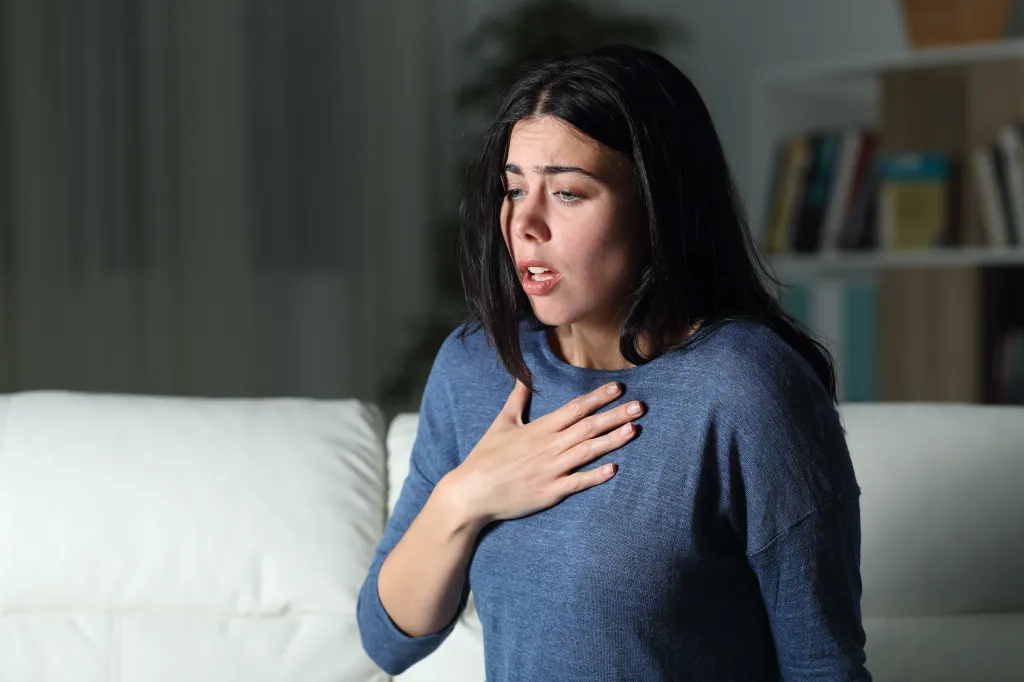 A woman with dark hair and a blue shirt sits on a white couch, clutching her chest and gasping for air.