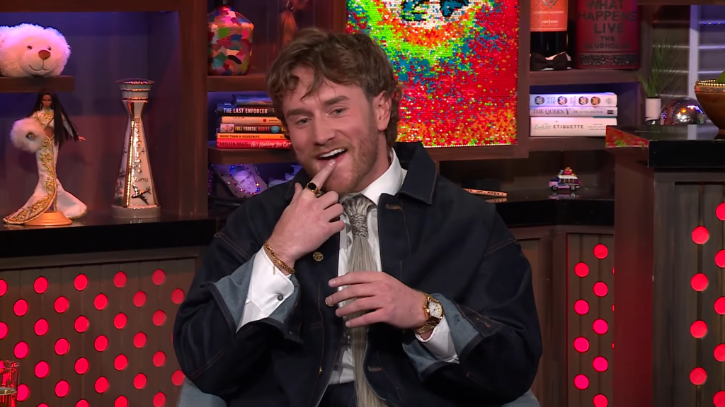 West Wilson smiling while sitting in a denim jacket and white shirt with a textured tie.