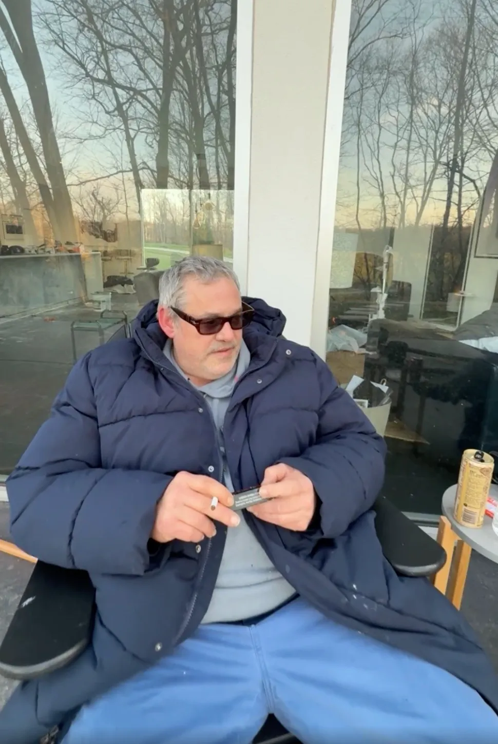 A man in sunglasses and a blue puffer jacket holding a cigarette and a small black box.