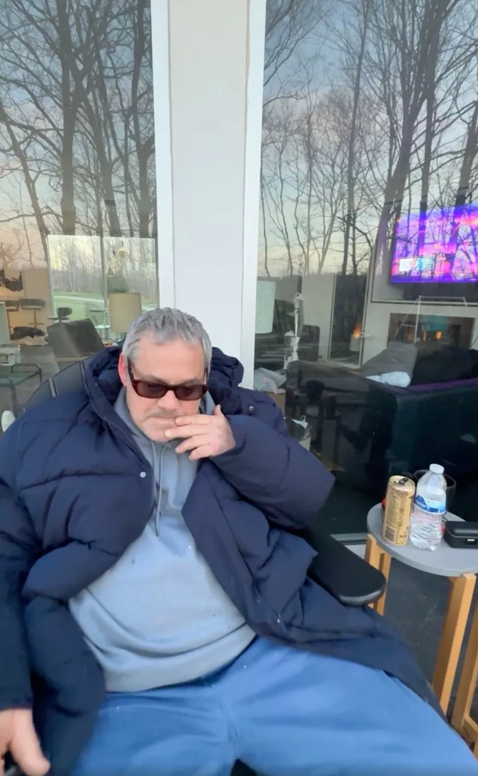 A man with gray hair and sunglasses sits in a chair wearing a puffer jacket, hoodie, and blue pants.