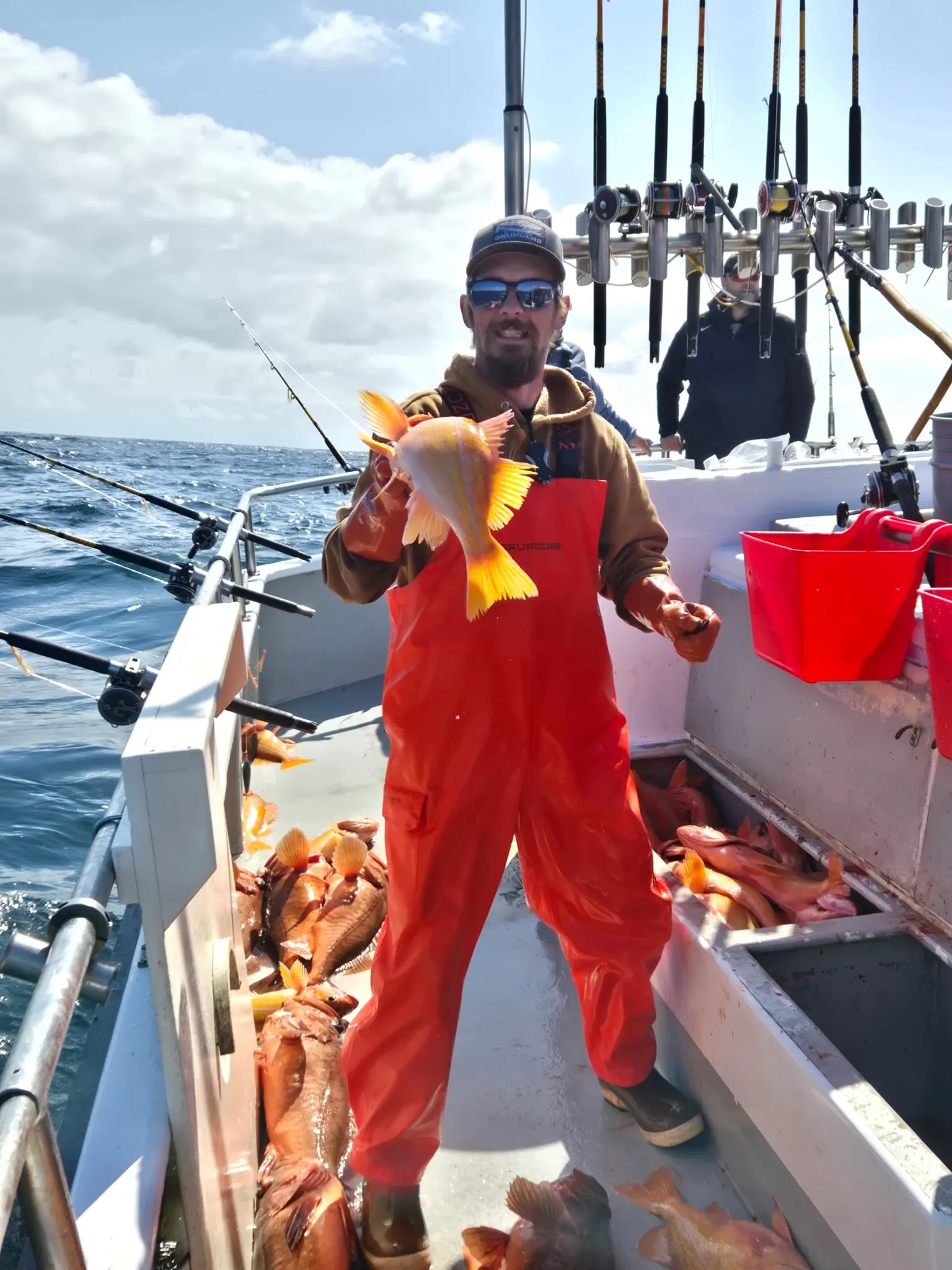A man in orange overalls on a boat holds up a fish as other fish lie in a bin beside him.