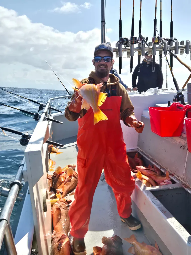 A man in orange overalls and sunglasses holds up a freshly caught fish on a boat filled with other fish.