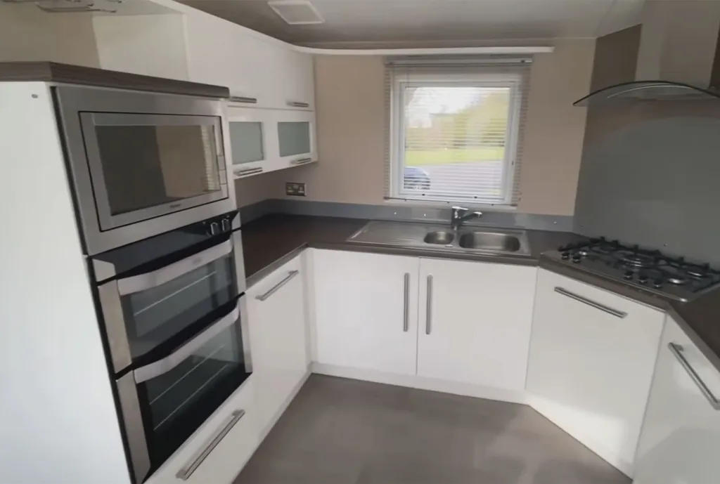 A clean, modern kitchen in a Willerby Meridian caravan with white cabinets, stainless steel appliances, and a window with blinds.